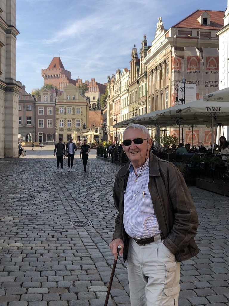 Stan Siedlecki standing in Old Market Square, Poznań, Poland — a sunny day with cobblestones, the historic facades of the square behind him, holding a walking stick